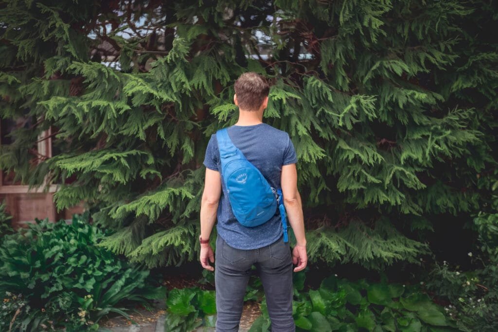 Best sling bag Osprey Daylite in serene park setting with lush greenery and pine trees.