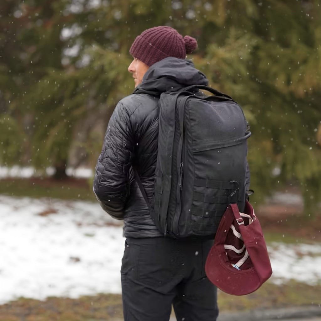 Goruck GR2 winter travel scene in snowy Detroit with black quilted jacket and evergreen backdrop.