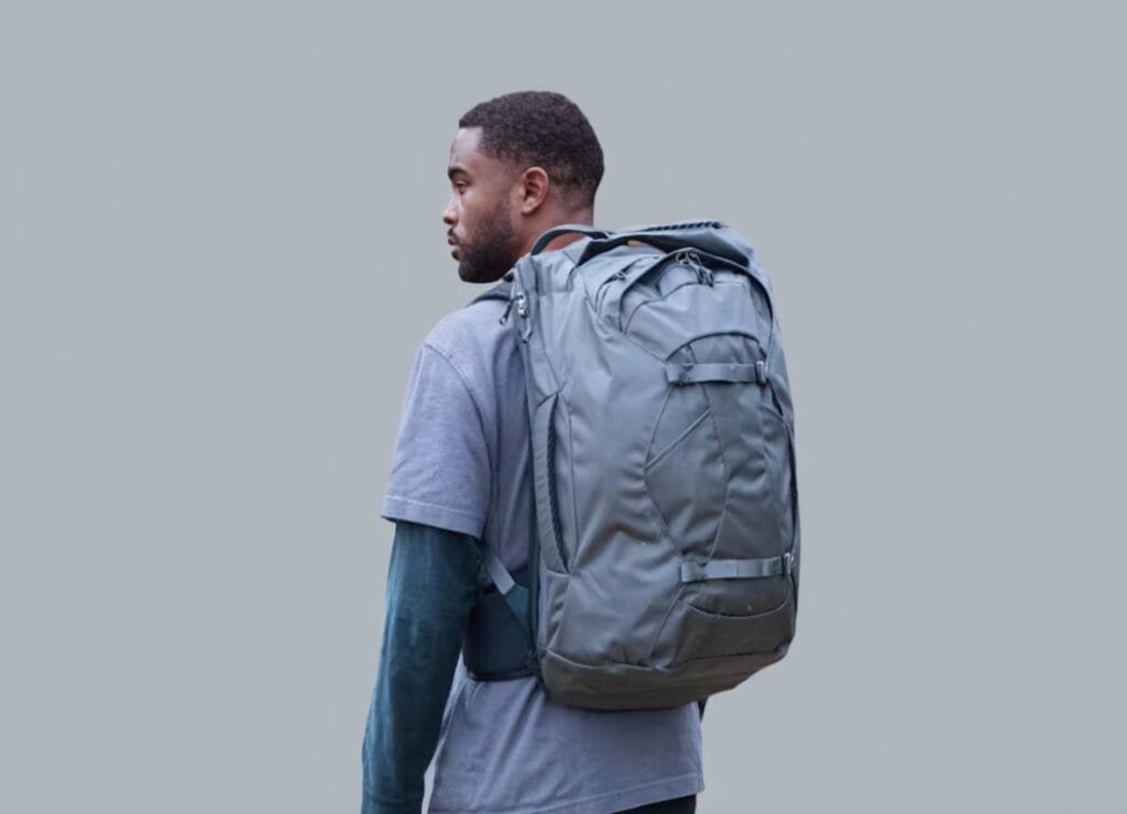 Osprey Farpoint 40 man wearing layered shirts with a utilitarian backpack against gray background.